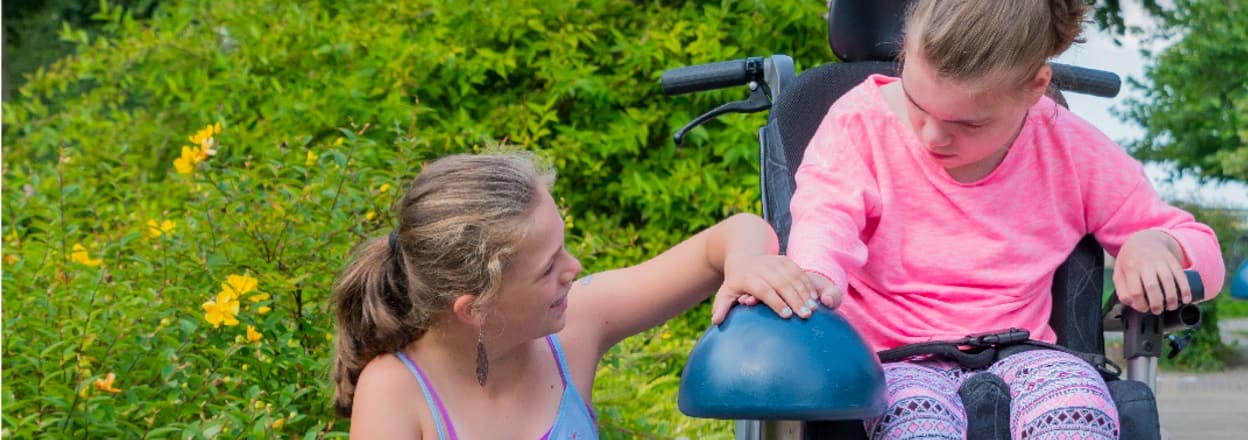 Photo of a young girl touching the hand of another young girl who is using a wheelchair